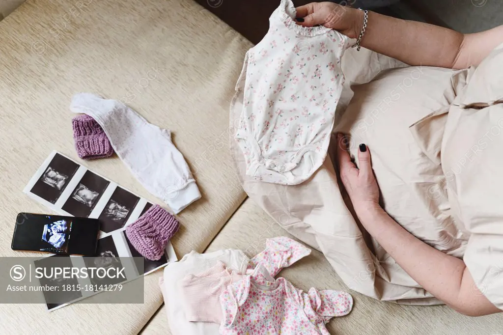 Pregnant woman with ultrasound scan holding baby cloths while sitting at home