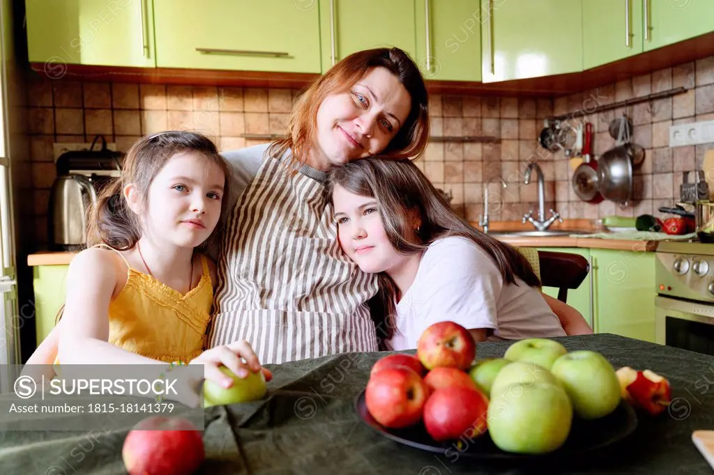 Smiling mother embracing daughters while sitting in kitchen at home