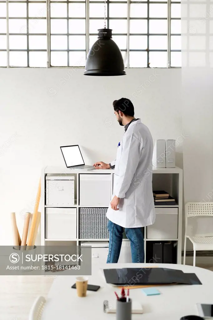 Male medical expert using laptop while standing at rack in hospital