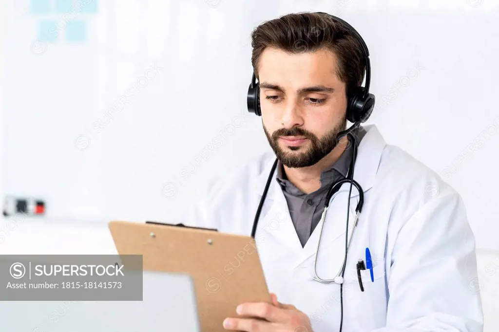 Handsome male doctor wearing headphone while reading report on clipboard in hospital