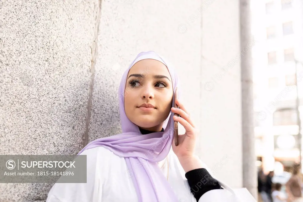 Arab teenage girl looking away while talking on smart phone by wall