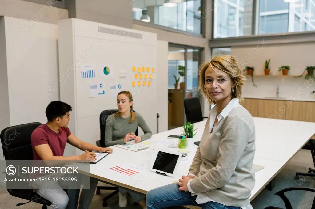 Businesswoman sitting on desk while male and female coworkers discussing in office