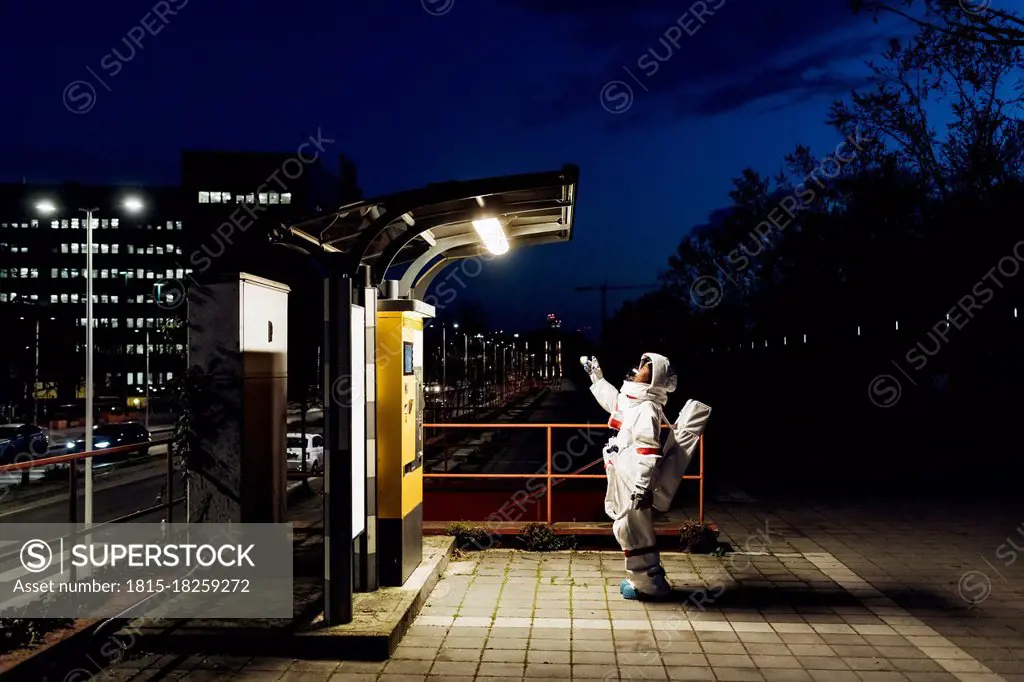 Young female astronaut standing near illuminated telephone booth in city during night