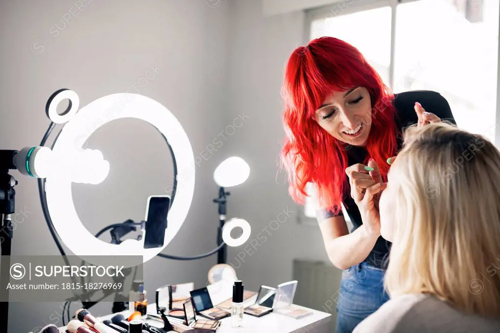 Smiling female make-up artist vlogging through smart phone while applying make-up to model at studio