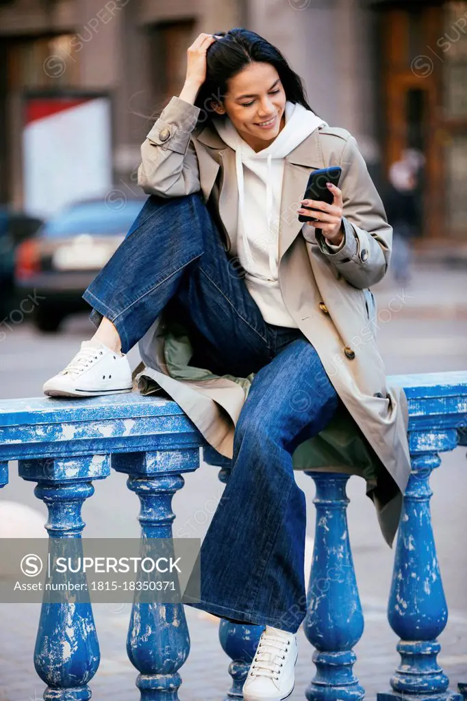 Smiling woman using mobile phone while sitting on blue railing
