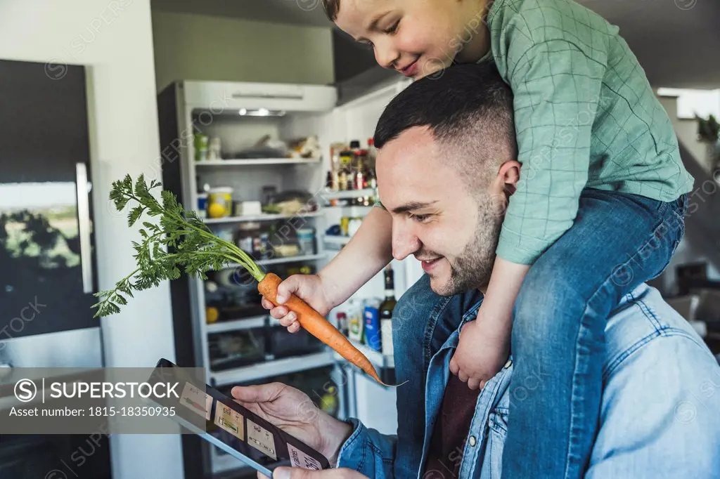Smiling man using tablet while son sitting on shoulder in kitchen at home