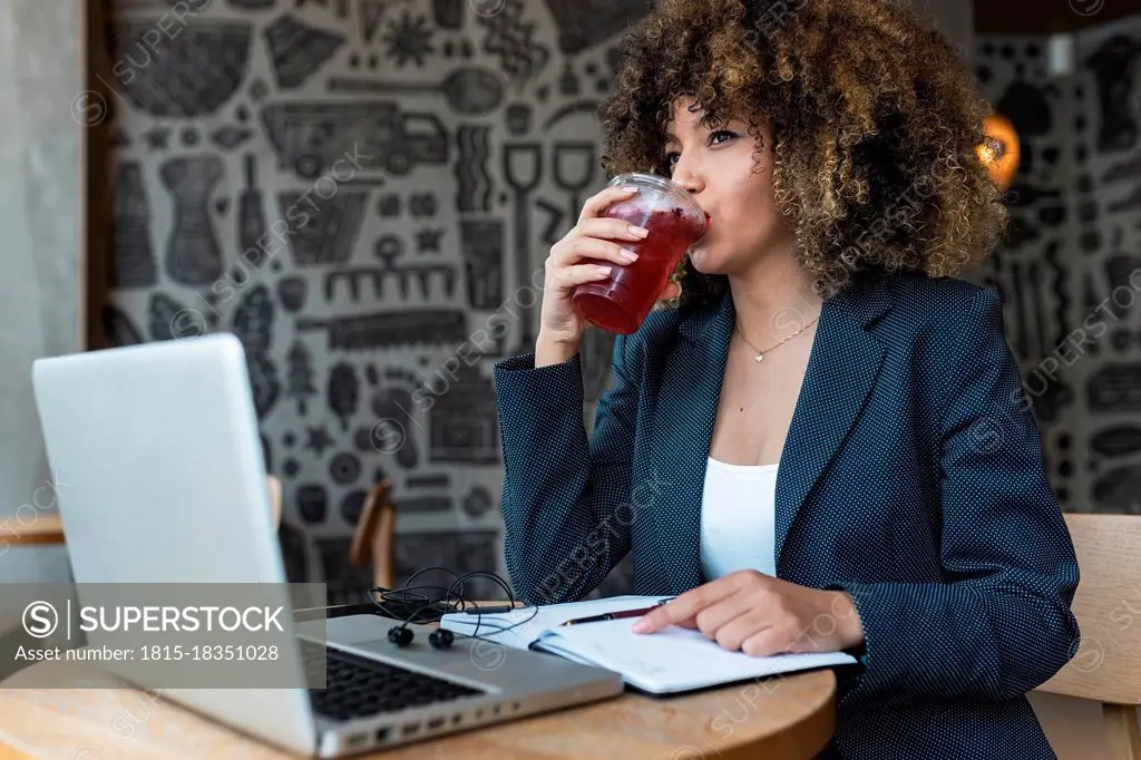 Afro businesswoman looking away while drinking in front of laptop