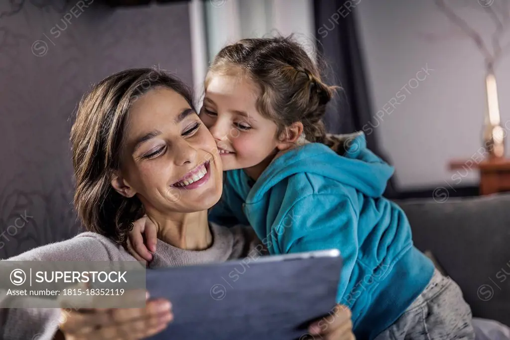 Girl kissing woman using digital tablet in living room