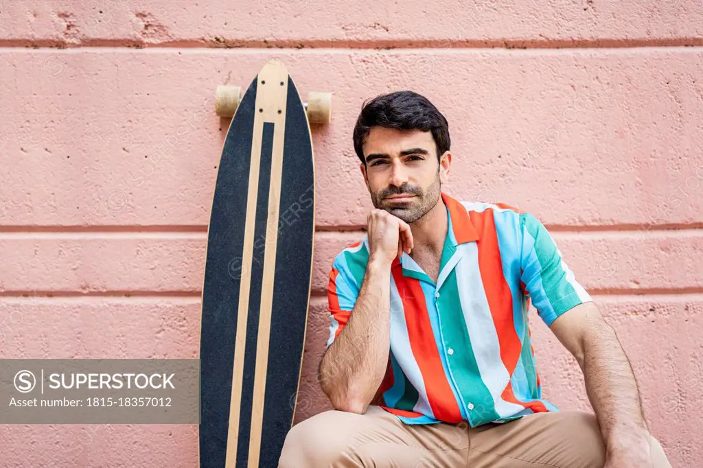 Handsome man with hand on chin sitting by longboard