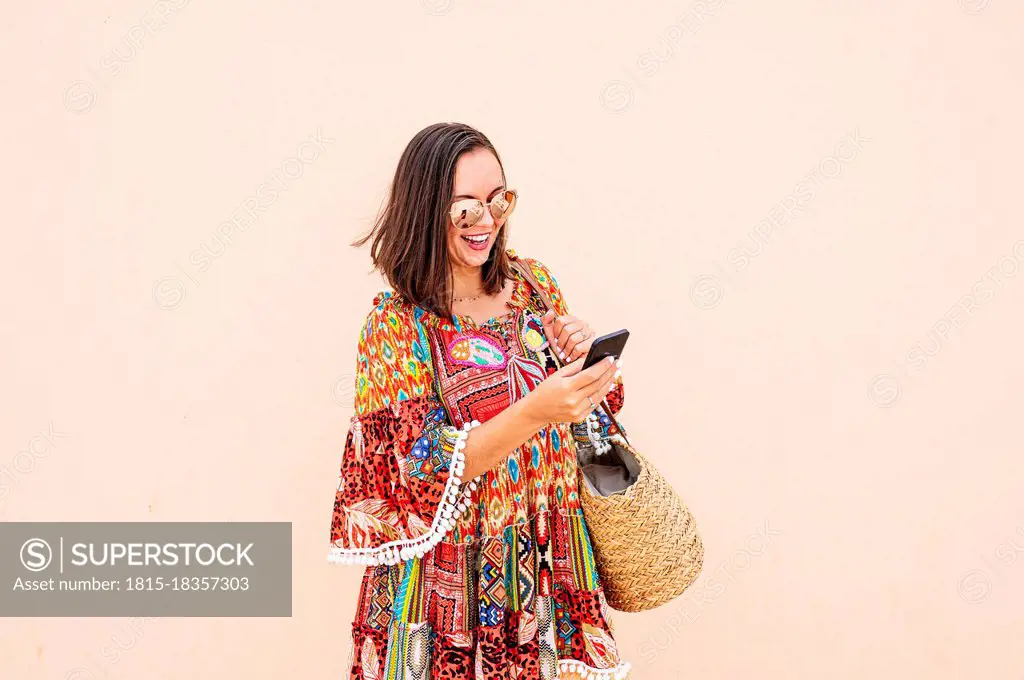 Cheerful woman wearing sunglasses using smart phone while standing in front of peach colored wall