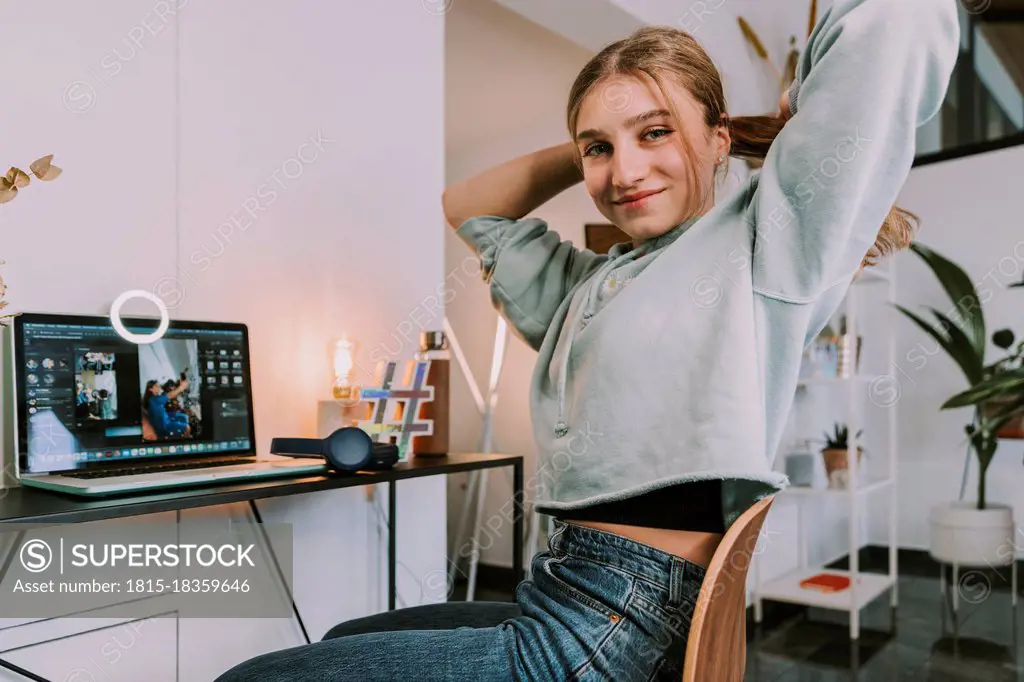 Smiling teenage female influencer with hand in hair sitting in front of laptop while vlogging at home