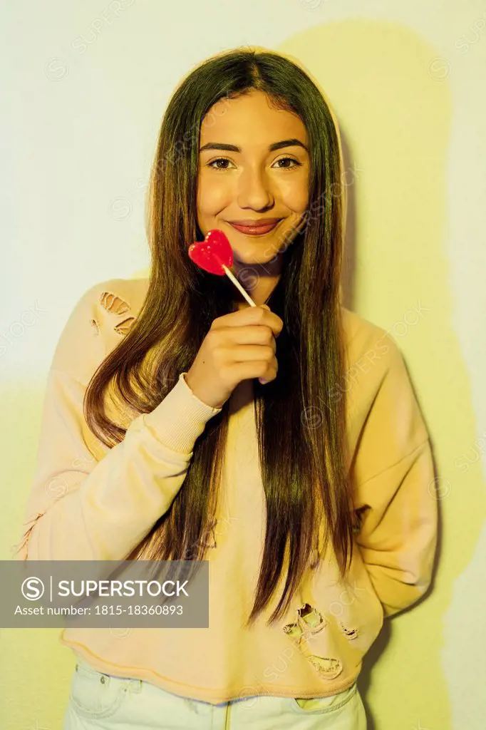 Beautiful woman holding heart shaped lollipop in front of wall