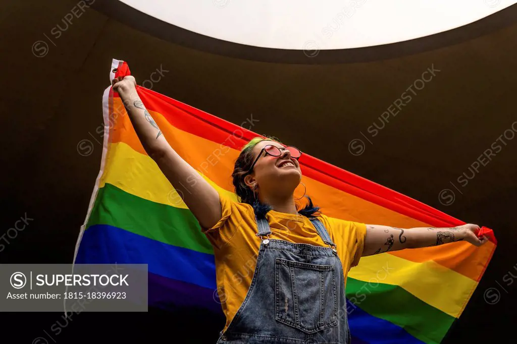 Smiling woman in bib overalls looking up while holding rainbow flag