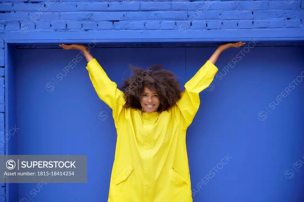 Smiling young Afro woman with arms raised in front of blue wall