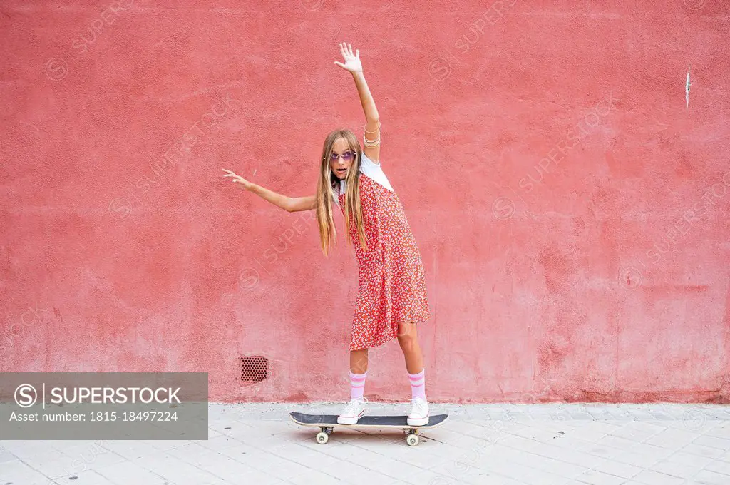 Pre-adolescent girl with hand raised skateboarding on footpath