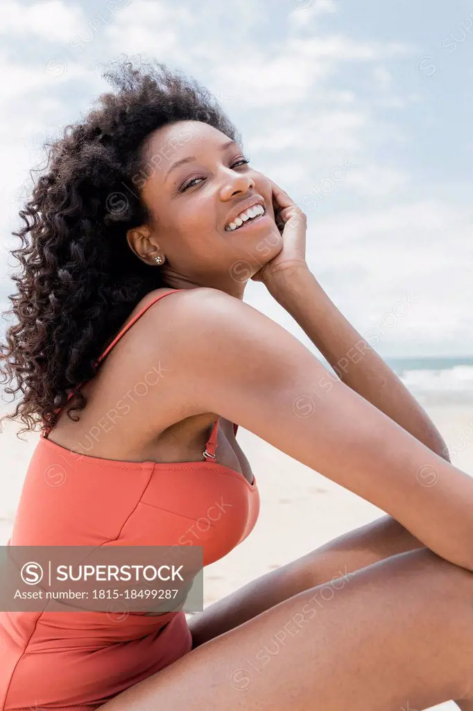 Smiling curly haired woman sitting with hand on chin at beach