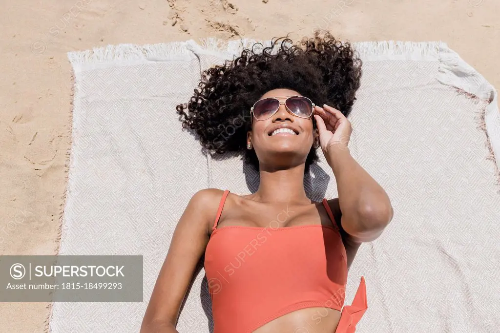 Smiling young woman with curly hair wearing sunglasses while lying on beach towel