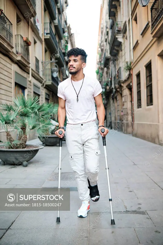Young man walking in alley with help of crutches amidst buildings