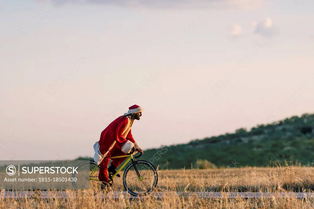 Young man in Santa Claus costume riding bicycle on road during sunset