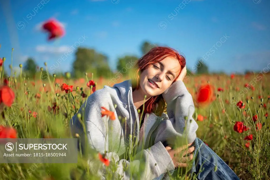 Redhead teenage girl sitting with eyes closed in poppy field on sunny day