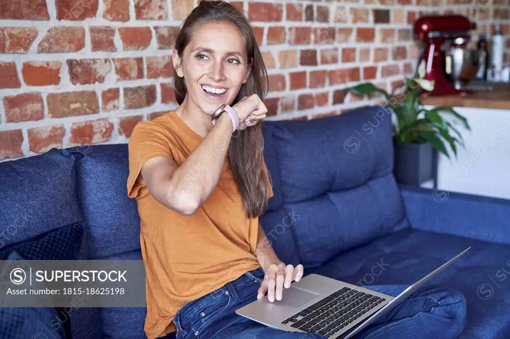 Woman with laptop talking through smart watch while sitting on sofa at home