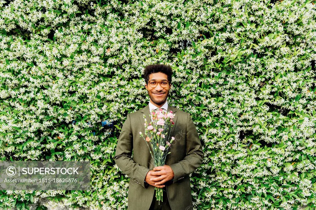 Happy businessman holding bouquet while standing in front of flowering plant