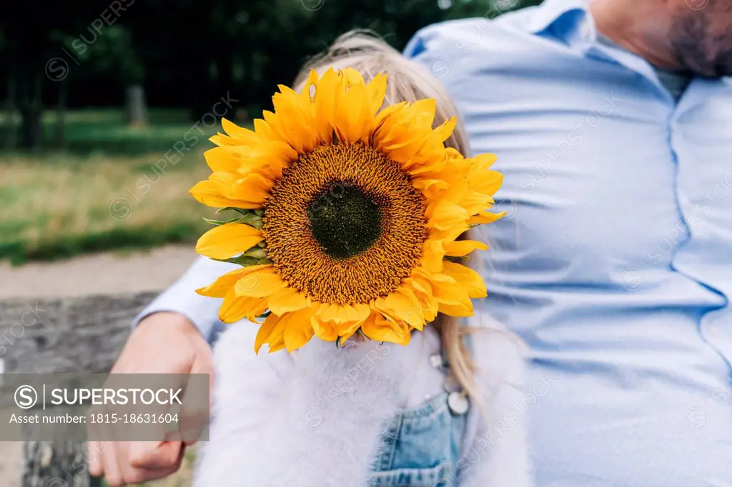 Girl covering face with sunflower while sitting by father at park