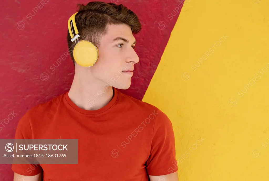 Young man listening music through wireless headphones in front of two tone wall