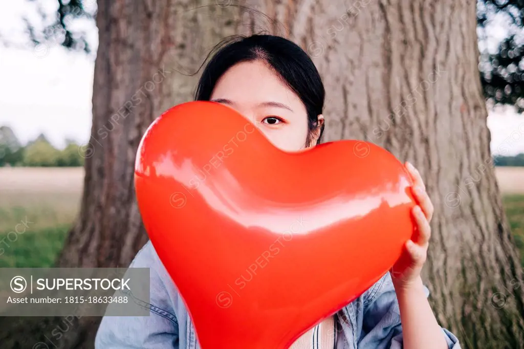 Woman covering face with red heart shaped balloon at park