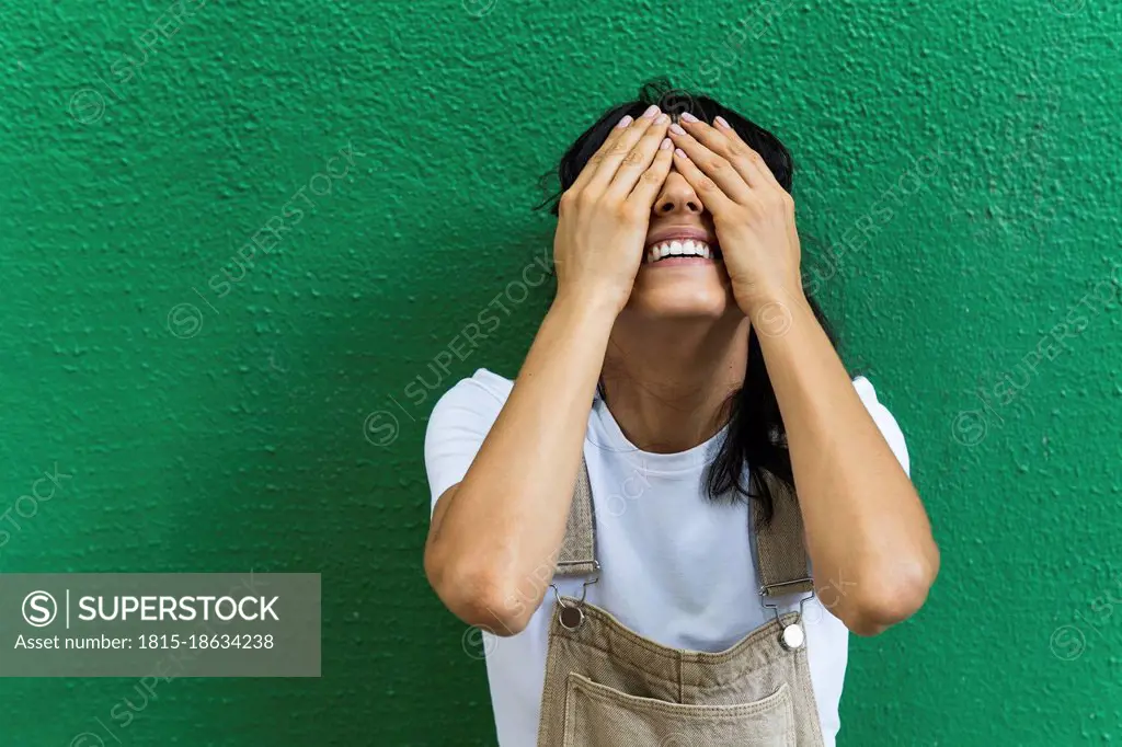 Smiling young woman covering eyes with hands in front of green wall