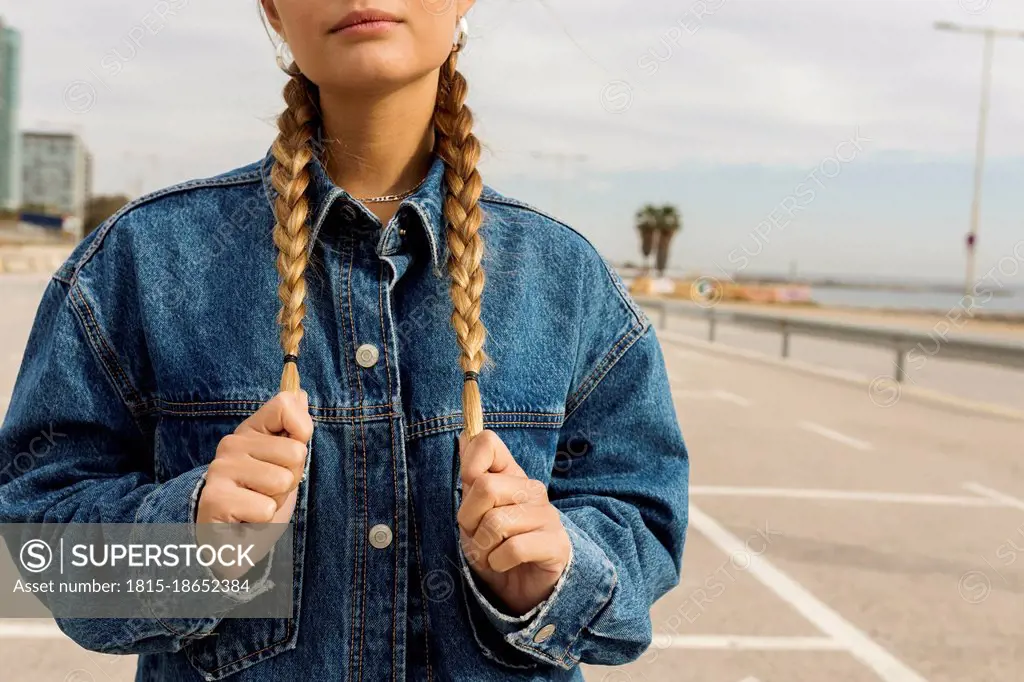 Young woman wearing denim jacket holding braided hair