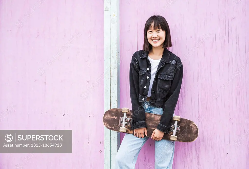 Happy teenage girl holding skateboard in front of wall