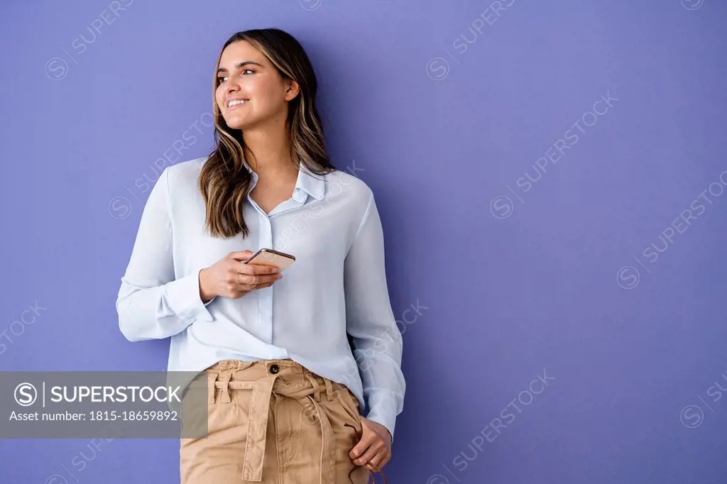 Female business professional holding smart phone while leaning on purple wall