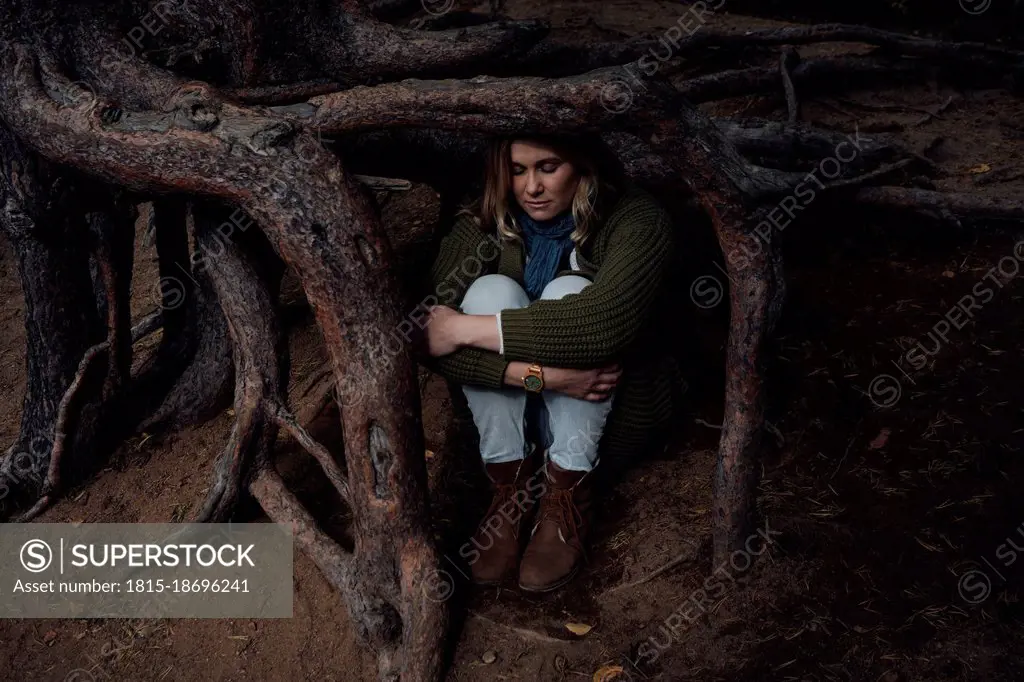 Lost woman sitting under tree roots while hugging knees