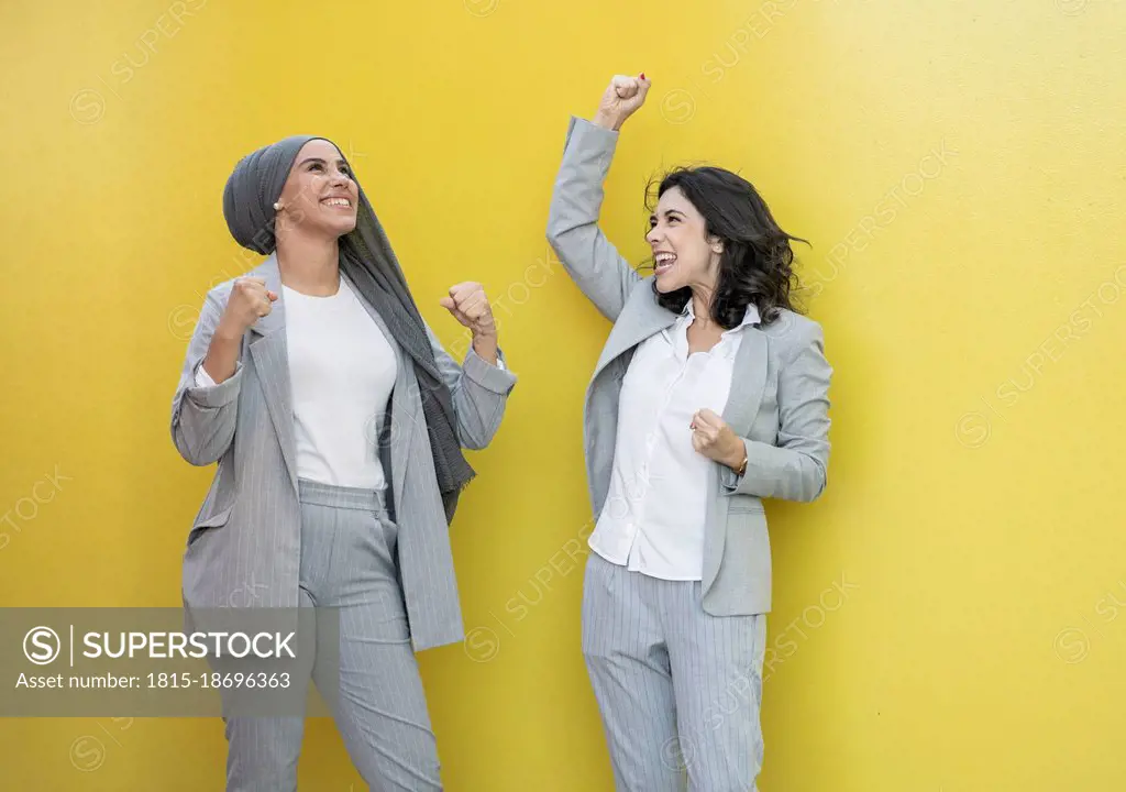 Cheerful female colleagues cheering in front of yellow wall