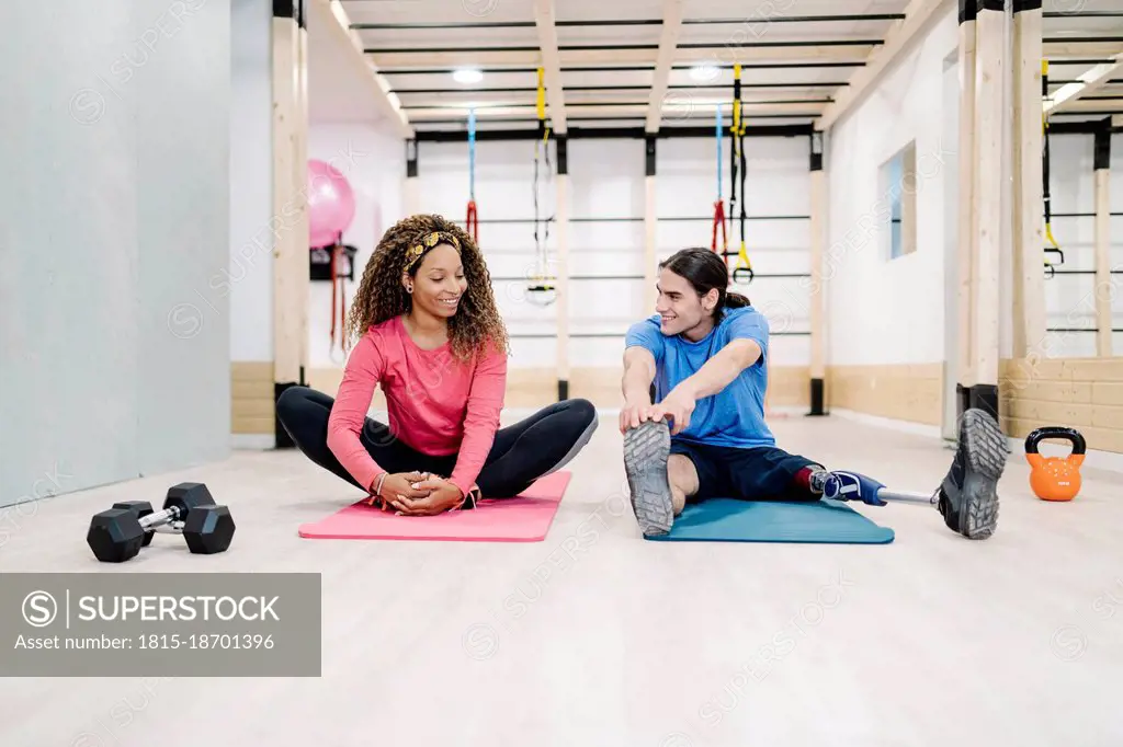 Smiling woman doing stretching exercise with disabled friend at gym