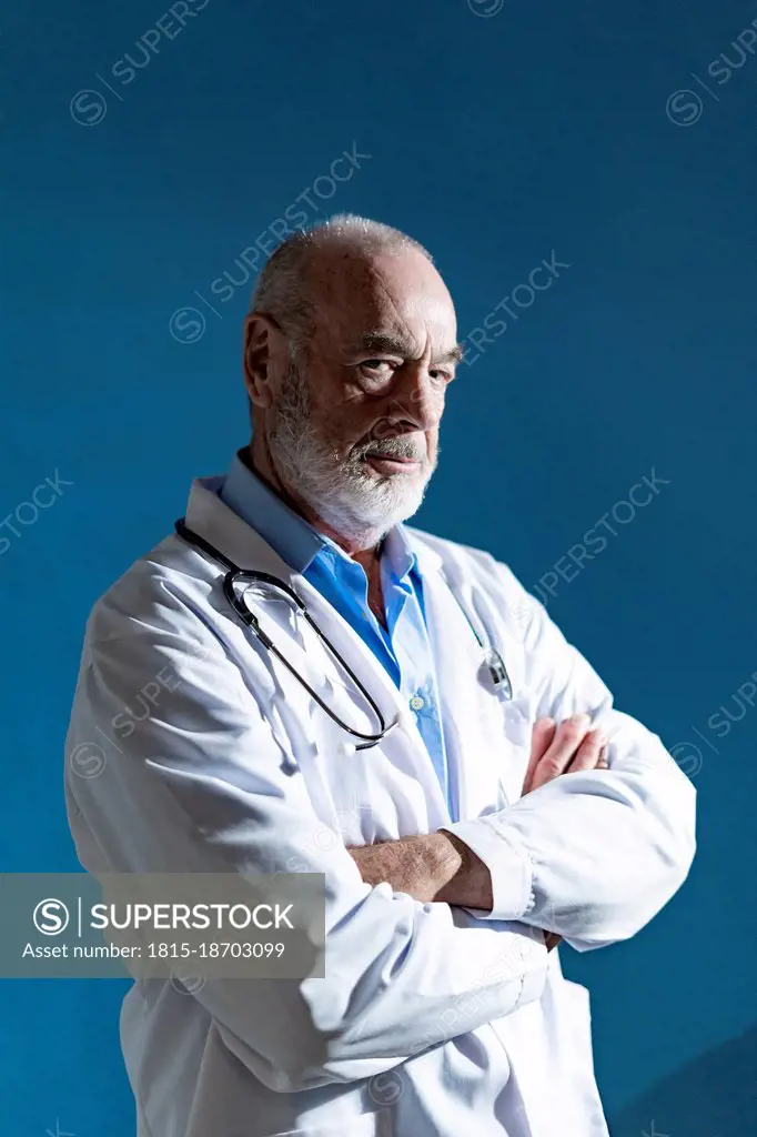 Senior doctor with arms crossed standing against blue background
