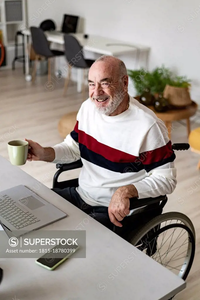Smiling businessman with coffee cup sitting on wheelchair at home