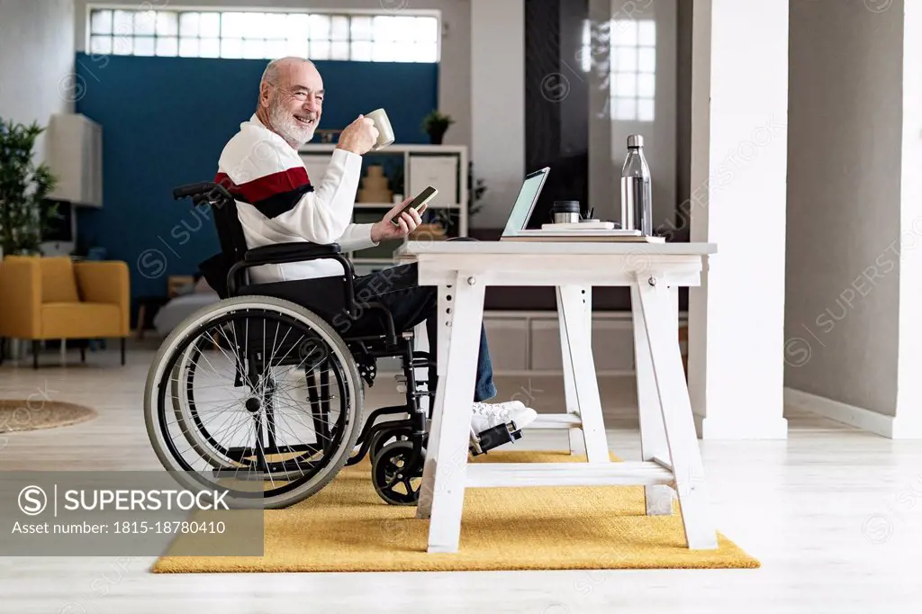 Smiling disabled freelancer holding coffee mug sitting at desk in home office
