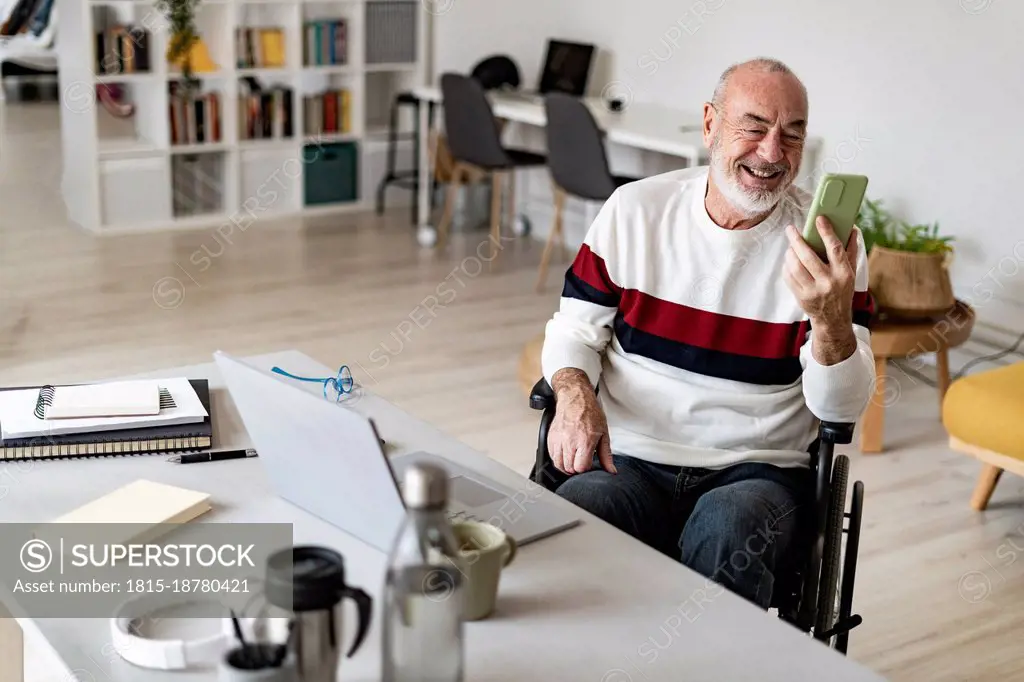 Senior businessman on wheelchair smiling on video call through smart phone at home office