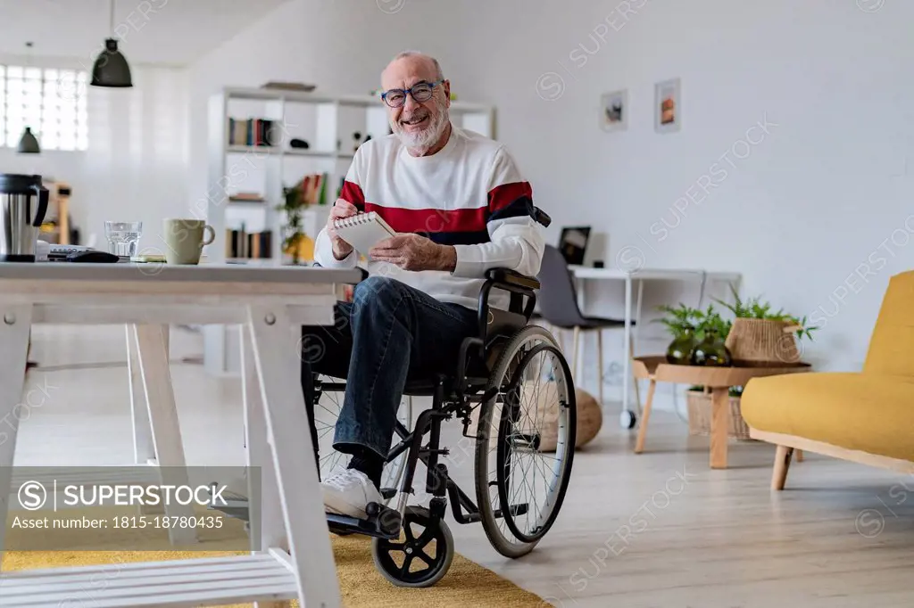 Smiling businessman sitting on wheelchair with notepad at home office