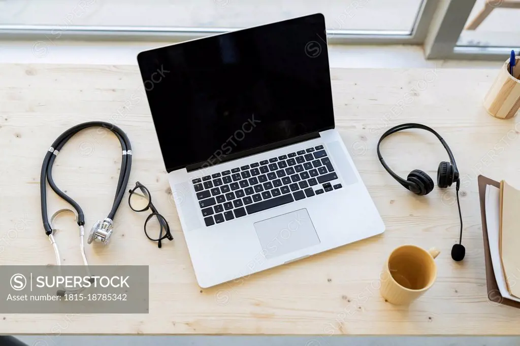 Laptop amidst stethoscope and mug with headphones on desk in home office