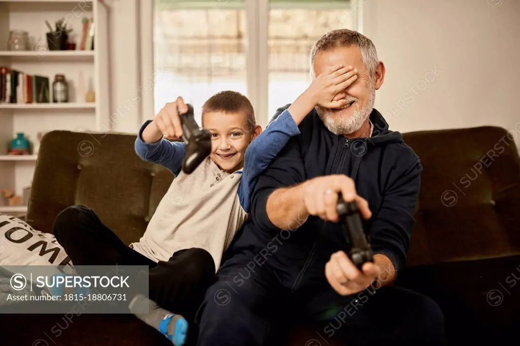 Grandson covering eyes of grandfather playing video game at home