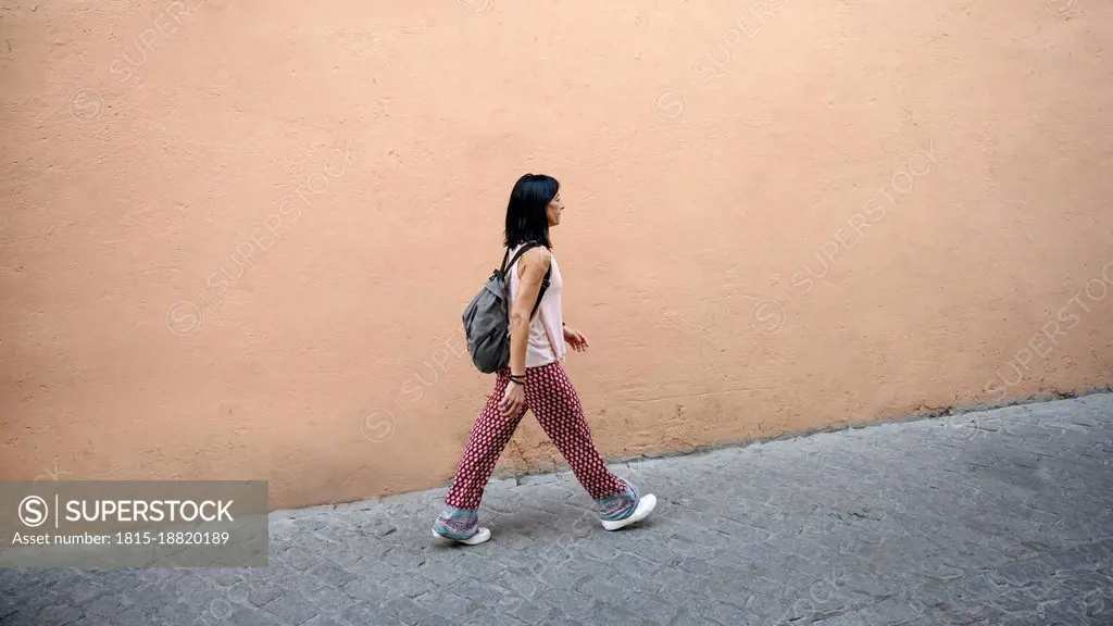 Woman with backpack waking on footpath by peach wall