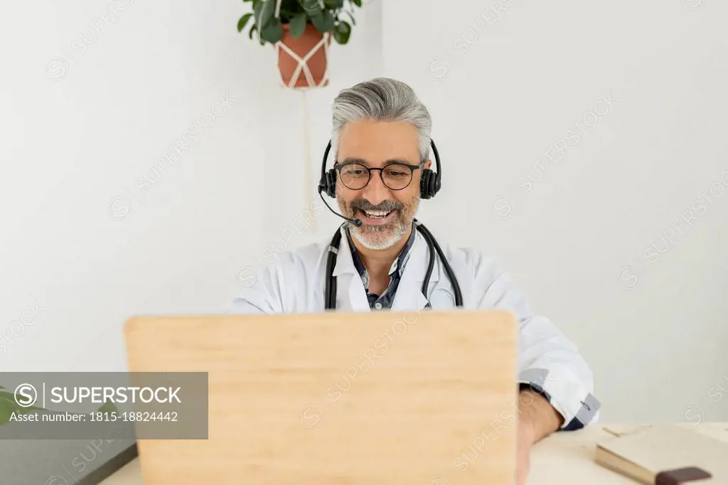 Smiling doctor with headphones on video call through laptop at home office