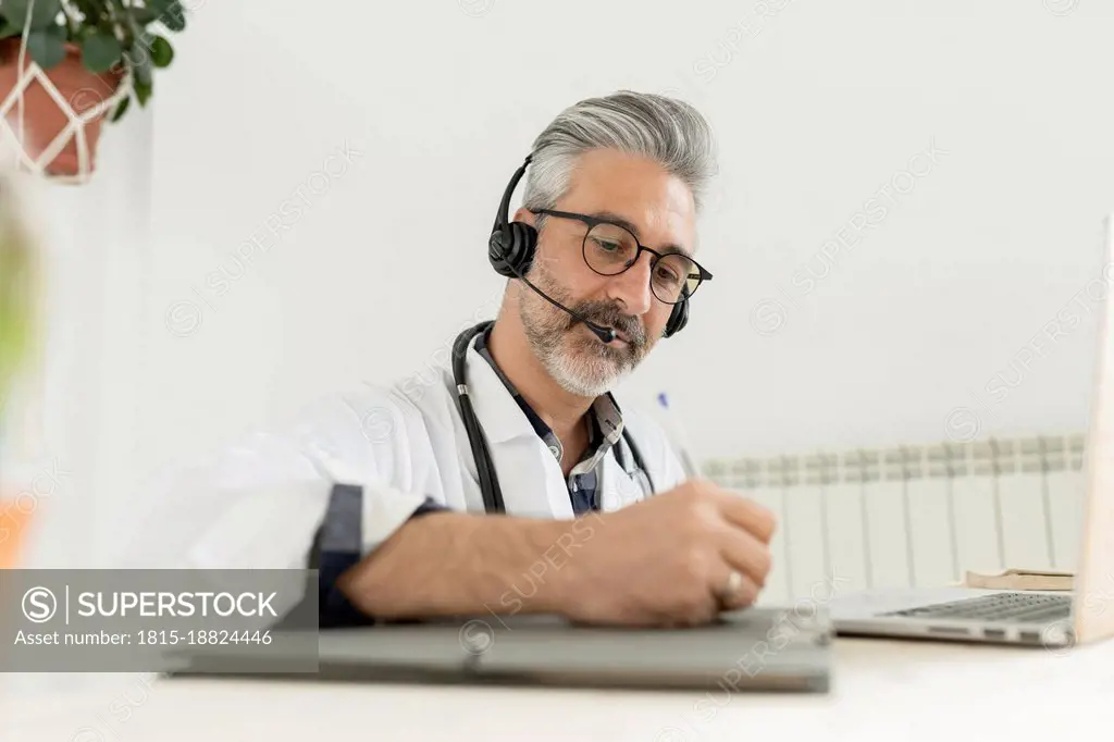 Doctor with headphones writing on clipboard at home office