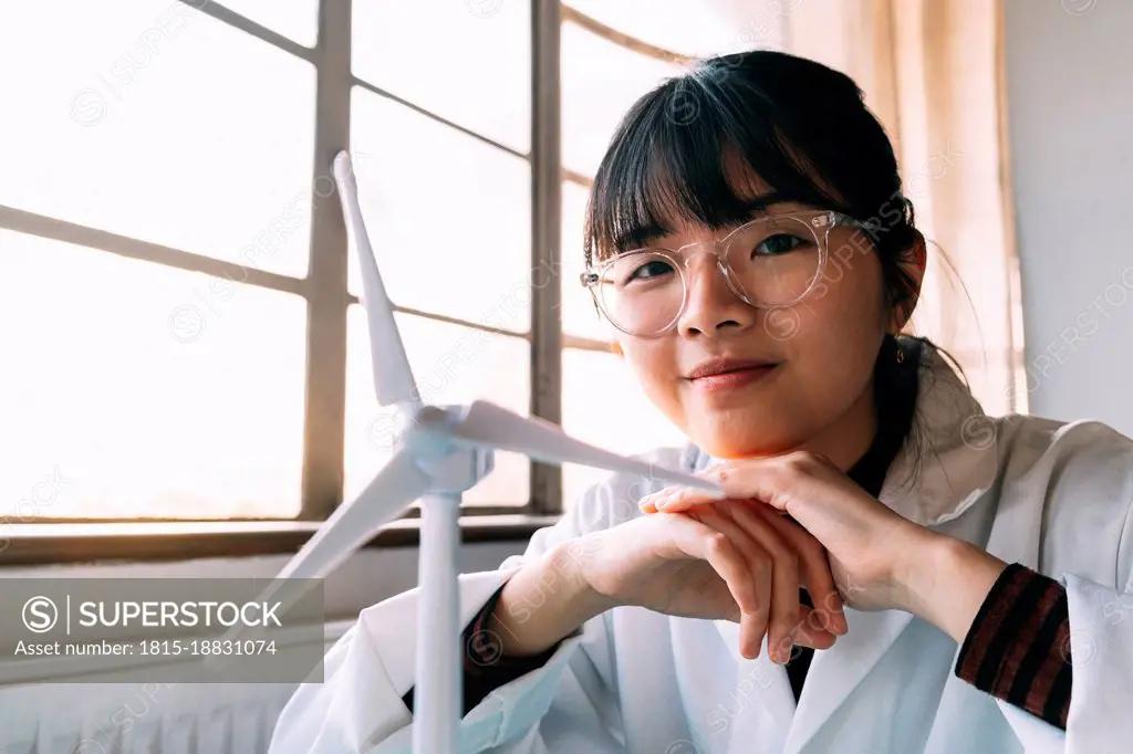Smiling young scientist sitting near wind turbine model in laboratory