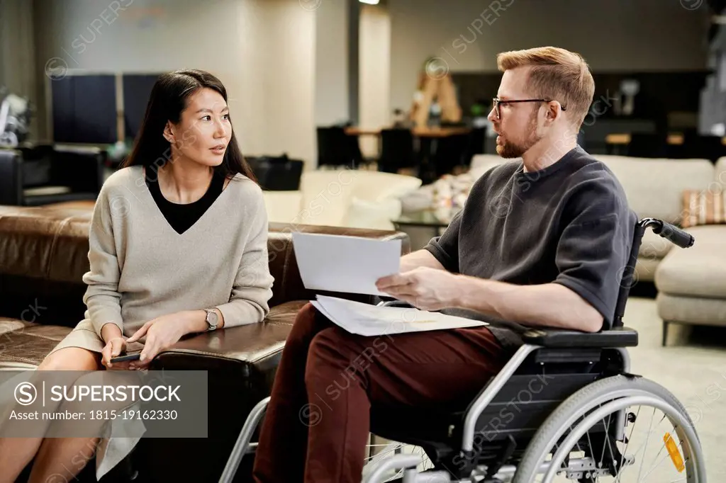 Disabled colleague with documents talking to businesswoman sitting on sofa in office