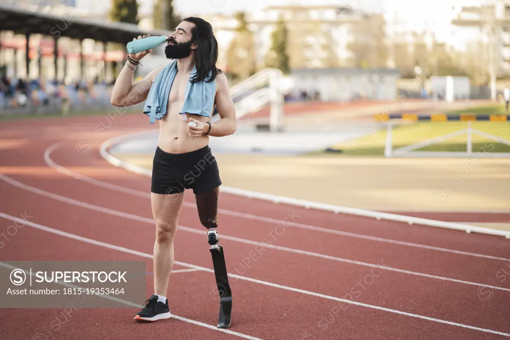 Athlete with prosthetic leg drinking water from bottle standing on running track