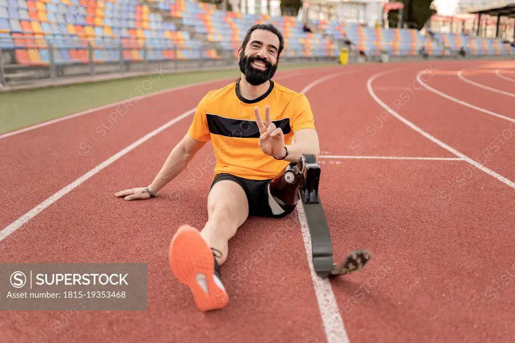Smiling man with prosthetic leg sitting on running track showing peace sign